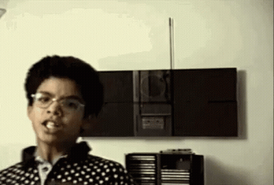 A Young Man Wearing Glasses Is Standing In Front Of A Stereo . GIF