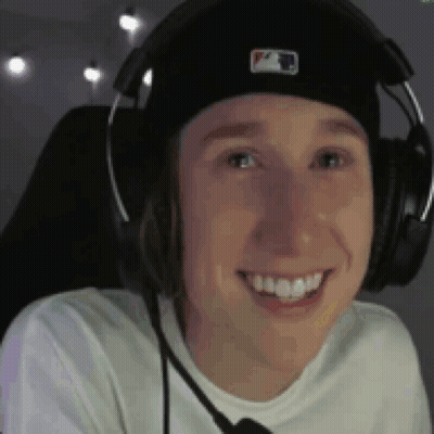A Young Man Wearing Headphones And A Hat Is Smiling While Sitting In A Chair . GIF