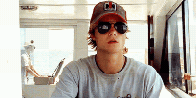 A Young Man Wearing Sunglasses And A Baseball Cap Is Sitting On A Boat . GIF