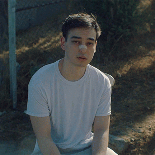 A Young Man With A Bandage On His Nose Sits On The Ground GIF
