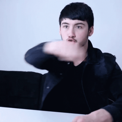 A Young Man With A Beard Is Sitting At A Table With His Arm Outstretched GIF