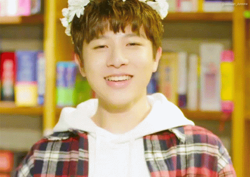 A Young Man With A Flower Crown On His Head Smiles In Front Of A Bookshelf GIF