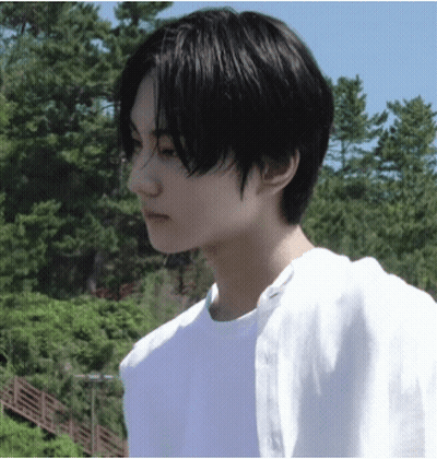 A Young Man With Black Hair And A White Shirt Is Standing In Front Of Trees GIF
