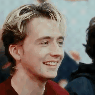 A Young Man With Blonde Hair And A Red Shirt Is Smiling And Looking At The Camera . GIF