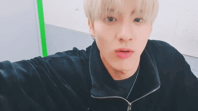 A Young Man With Blonde Hair Is Taking A Selfie While Wearing A Black Jacket And Necklace . GIF