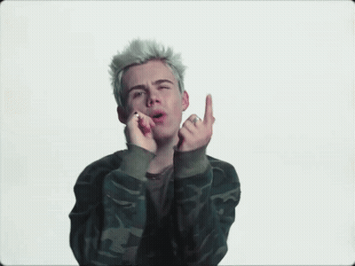 A Young Man With Blonde Hair Is Wearing A Camouflage Jacket And Giving The Middle Finger . GIF