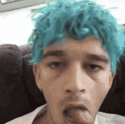 A Young Man With Blue Hair Is Sticking His Tongue Out While Sitting On A Couch . GIF