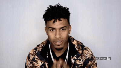 A Young Man With Curly Hair And A Beard Is Wearing A Floral Jacket And Necklace . GIF