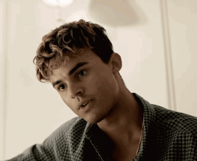 A Young Man With Curly Hair And A Plaid Shirt Looks At The Camera GIF