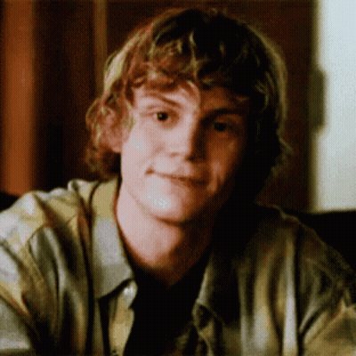 A Young Man With Curly Hair Is Sitting At A Table Looking At The Camera . GIF