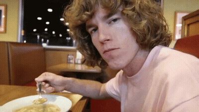 A Young Man With Curly Hair Is Sitting At A Table With A Plate Of Food And A Fork In His Hand . GIF