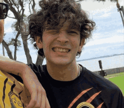 A Young Man With Curly Hair Is Smiling For The Camera While Standing Next To Another Man . GIF