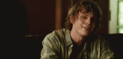 A Young Man With Curly Hair Is Smiling In A Dark Room . GIF
