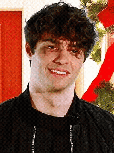 A Young Man With Curly Hair Is Smiling In Front Of A Red Christmas Tree GIF