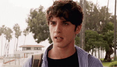 A Young Man With Curly Hair Is Standing In Front Of A Lifeguard Tower And Palm Trees . GIF
