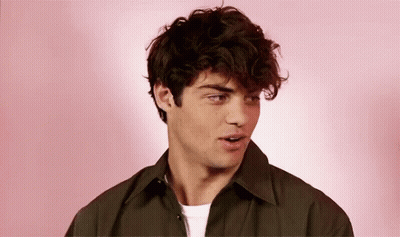 A Young Man With Curly Hair Is Standing In Front Of A Pink Background And Looking At The Camera . GIF
