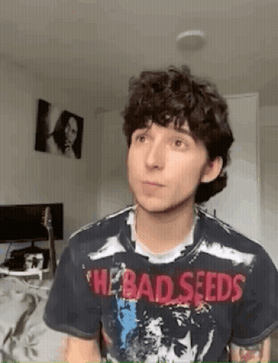 A Young Man With Curly Hair Is Wearing A Bad Seeds T-Shirt And Looking At The Camera . GIF