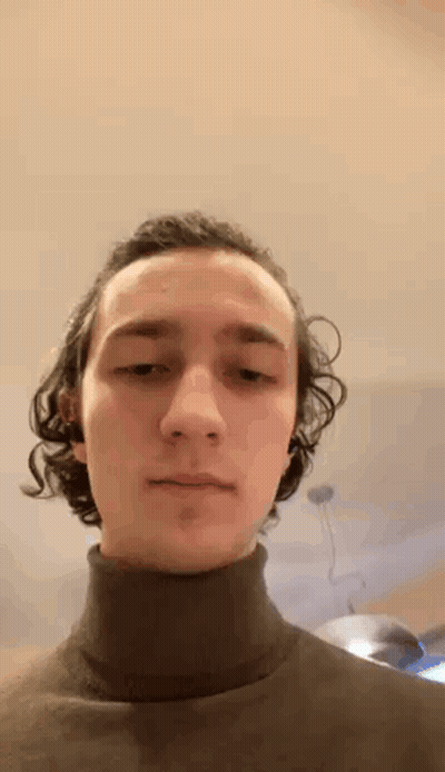 A Young Man With Curly Hair Is Wearing A Black Turtleneck Sweater And Looking At The Camera . GIF