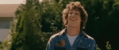 A Young Man With Curly Hair Is Wearing A Blue Shirt And Smiling In Front Of Trees . GIF
