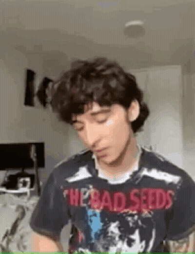 A Young Man With Curly Hair Is Wearing A T-Shirt With The Bad Seeds On It . GIF