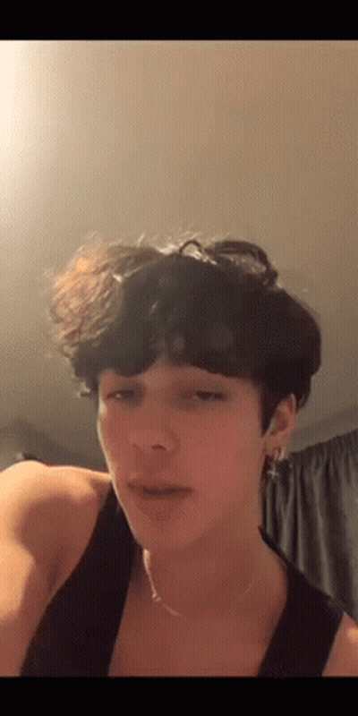 A Young Man With Curly Hair Is Wearing A Tank Top And Earrings . GIF