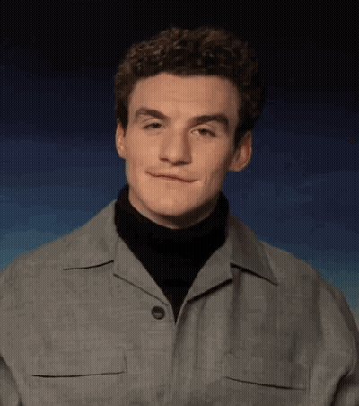 A Young Man With Curly Hair Is Wearing A Turtleneck And A Grey Jacket . GIF