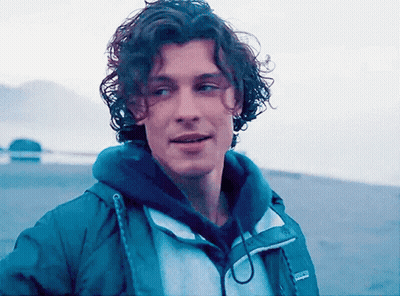 A Young Man With Curly Hair Wearing A Blue Jacket And A Hoodie Is Standing On A Beach . GIF