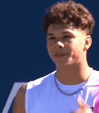 A Young Man With Curly Hair Wearing A White Tank Top And A Necklace Is Smiling . GIF