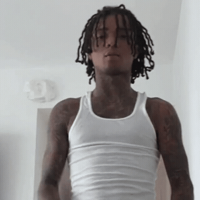 A Young Man With Dreadlocks Is Wearing A White Tank Top And Standing In A Room . GIF