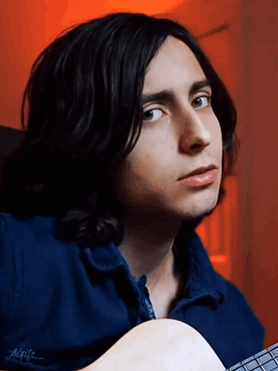 A Young Man With Long Black Hair Is Holding A Guitar And Wearing A Blue Jacket GIF