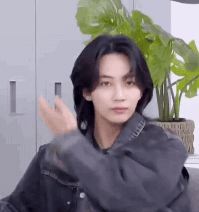 A Young Man With Long Black Hair Is Standing In Front Of A Potted Plant In A Room . GIF
