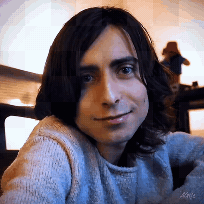 A Young Man With Long Dark Hair Is Wearing A Sweater And Smiling . GIF