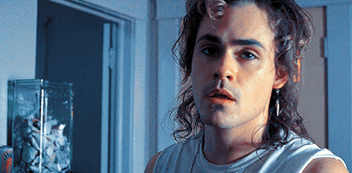 A Young Man With Long Hair And Earrings Is Standing In Front Of A Door . GIF