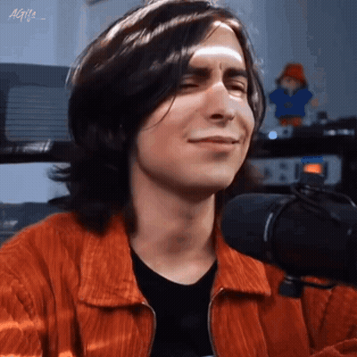 A Young Man With Long Hair Is Smiling In Front Of A Microphone . GIF