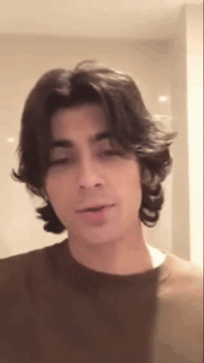A Young Man With Long Hair Is Wearing A Brown Shirt And Talking . GIF