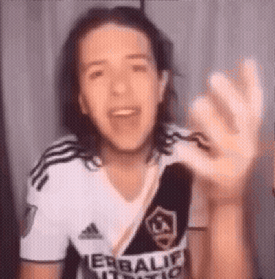 A Young Man With Long Hair Is Wearing A White And Black Adidas Shirt And Waving His Hand . GIF