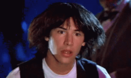 A Young Man With Long Hair Is Wearing A White Shirt And A Black Vest And Looking At The Camera . GIF