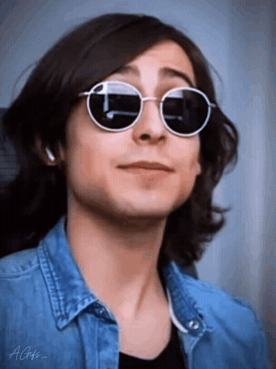A Young Man With Long Hair Is Wearing Sunglasses And A Denim Shirt . GIF