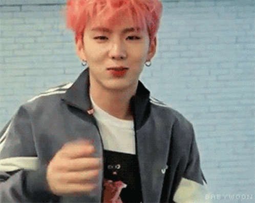 A Young Man With Pink Hair And A Gray Jacket Is Standing In Front Of A White Brick Wall . GIF