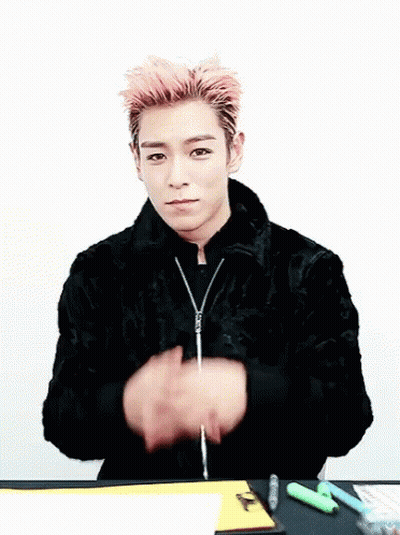 A Young Man With Pink Hair Is Sitting At A Table Making A Heart With His Hands GIF