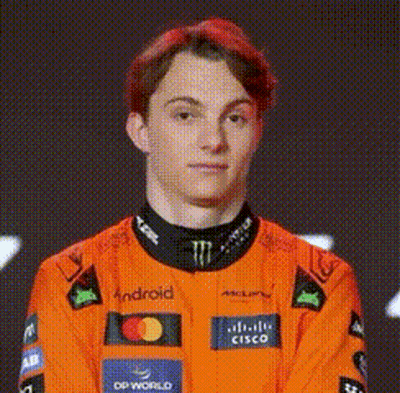A Young Man With Red Hair Is Wearing An Orange Racing Suit And Looking At The Camera . GIF