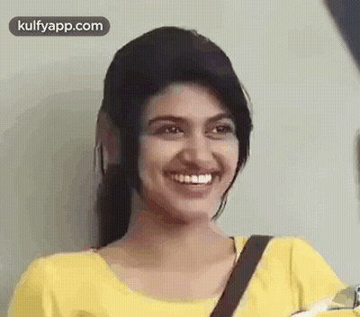A Young Woman In A Yellow Shirt Is Smiling And Looking At The Camera . GIF