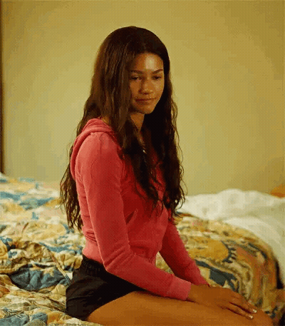 A Young Woman Is Sitting On A Bed Wearing Shorts And A Pink Sweater . GIF