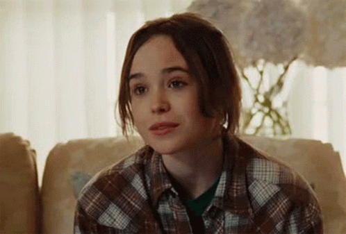 A Young Woman Is Sitting On A Couch Wearing A Plaid Shirt And Looking At The Camera . GIF