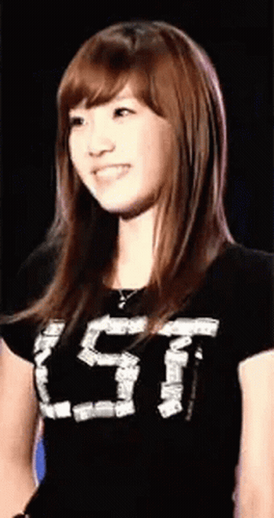 A Young Woman Wearing A Black T-Shirt With The Letter T On It Is Smiling And Looking At The Camera . GIF