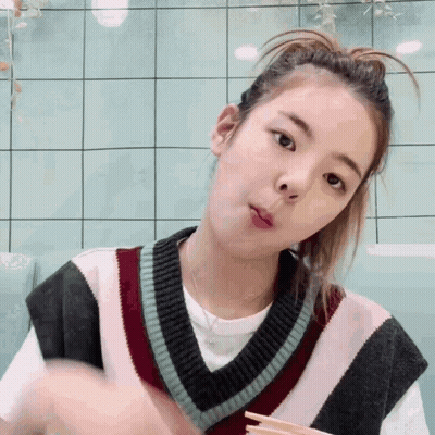 A Young Woman Wearing A Sweater Vest And A Ponytail Is Eating With Chopsticks . GIF