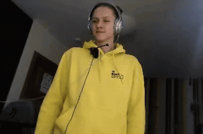 A Young Woman Wearing A Yellow Hoodie And Headphones Is Standing In A Room . GIF