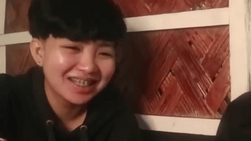 A Young Woman With Braces On Her Teeth Is Smiling While Sitting In Front Of A Wooden Wall . GIF