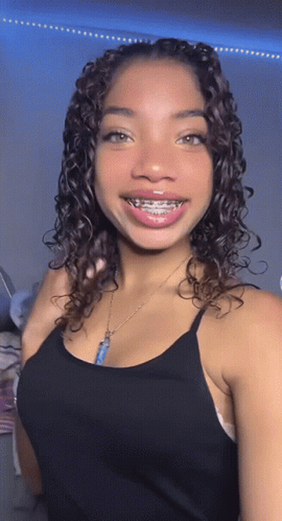 A Young Woman With Curly Hair And Braces On Her Teeth Is Wearing A Black Tank Top And A Necklace . GIF