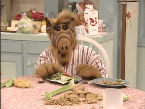 Alf From The Tv Show Sits At A Table Eating A Plate Of Food GIF